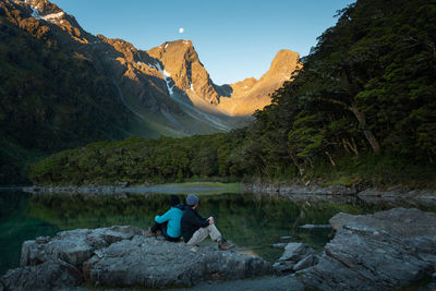 Man sitting on rock by lake against mountain