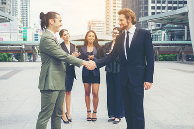 Colleagues clapping while happy coworkers handshaking on footpath against buildings in city