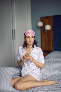 Woman in pajamas and with a sleep mask on the bed in the bedroom and drinking orange juice 