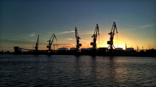 Silhouette cranes at commercial dock against sky during sunset