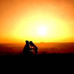 Silhouette couple sitting on landscape against orange sunset sky