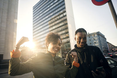 Portrait of smiling teenage girl flexing muscles while standing with friend in city