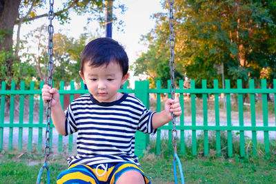 Cute boy sitting on swing against fence 