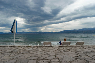 Rear view of woman reading book while sitting on deck chair at beach against cloudy sky