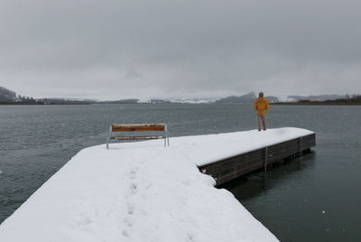 Rear view of man standing at lake against sky during snowfall