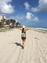 Rear view of young woman with backpack and hat walking at beach against blue sky