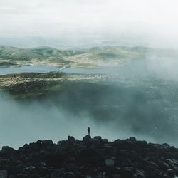 Person standing on rocks by landscape against sky during foggy weather