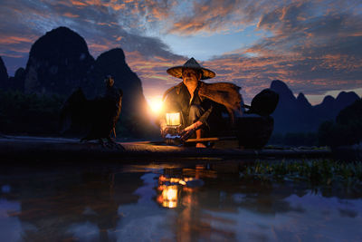 Man sitting by lake against sky during sunset