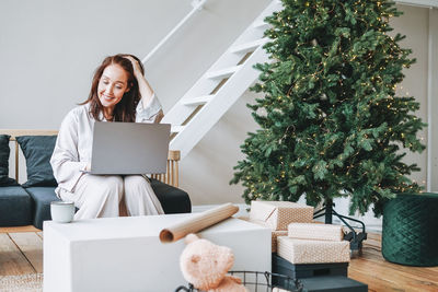 Young adult forty years woman with present gift box using laptop in living room with christmas tree 
