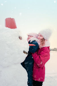 Teenage girl carrying sister by snowman against sky