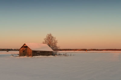 House on snow covered land against sky during sunset