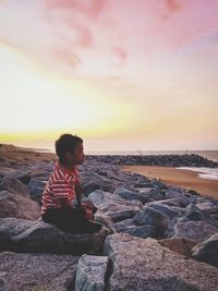 Side view of child sitting on rock at beach against sky during sunset