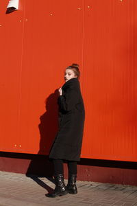 Fashionable teenage girl wearing black warm clothing standing against red wall during sunny day