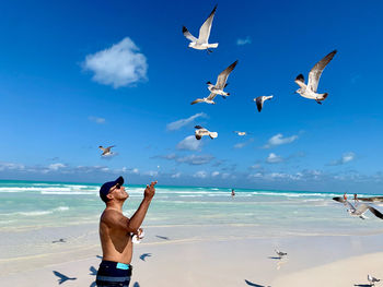 Shirtless man feeding birds at beach against sky