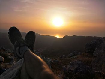 Low section of man relaxing on rock against sky during sunset