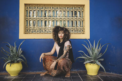 Full length portrait of woman crouching amidst potted cacti by blue wall