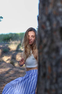 Young, blonde and beautiful woman hiding behind a tree covering half of her body in a pine forest.