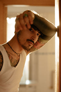 Side view of young stylish guy with mustache and tattoos wearing white tank top and leaning against wall while looking at camera