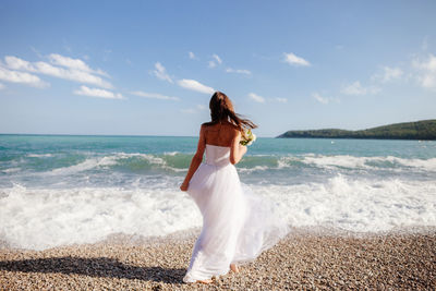 Rear view of bride holding flowers at beach on sunny day