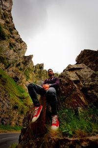 Low angle view of man sitting on rock formation against sky at cheddar gorge