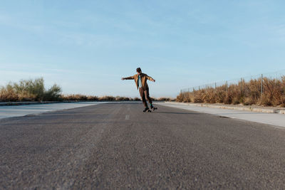 Full body young bearded male skater in stylish wear riding skateboard along asphalt road in countryside