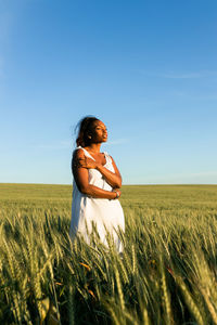 Young black lady in white summer dress strolling on green wheat field while looking away in daytime under blue sky