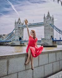 Full length of woman sitting on retaining wall against tower bridge in city