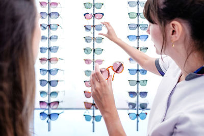 Side view of female doctor helping female client picking up eyeglasses from rack in modern optic office