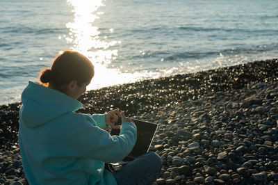 Woman sitting with laptop by sea on sunny day