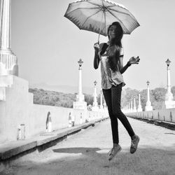 Portrait of woman with umbrella jumping over footpath against sky