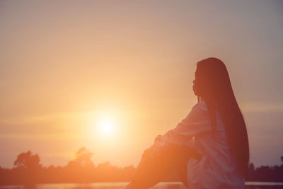 Side view of woman sitting against orange sky