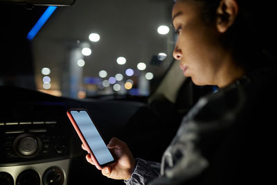 Side view of focused female passenger browsing mobile phone with blank screen in city at night