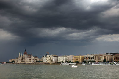 Buildings by danube river against cloudy sky in city