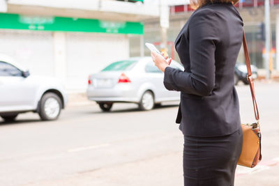 Midsection of businesswoman holding mobile phone while standing on road in city
