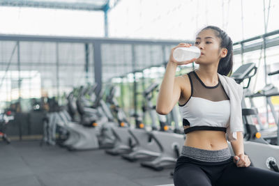 Young woman drinking water from bottle while sitting at gym