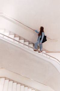 From above side view of faceless distant female with long hair in elegant wear with handbag walking upstairs in corridor of light spacious building