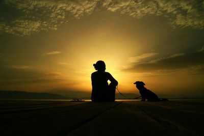 Rear view of silhouette woman with dog relaxing on pier in front sea at sunset