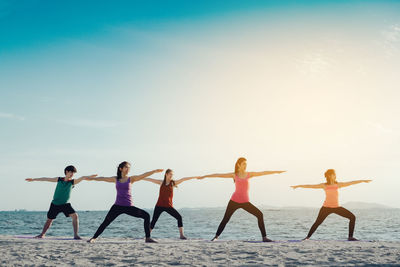 Young friends practicing yoga in warrior position on shore at beach during sunny day