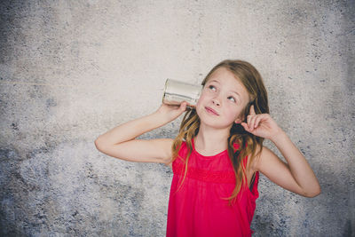 Cute girl playing with tin can phone while standing against wall