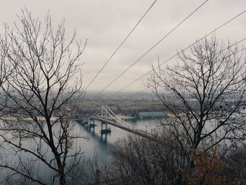 Bare trees blocking view of bridge over river against sky