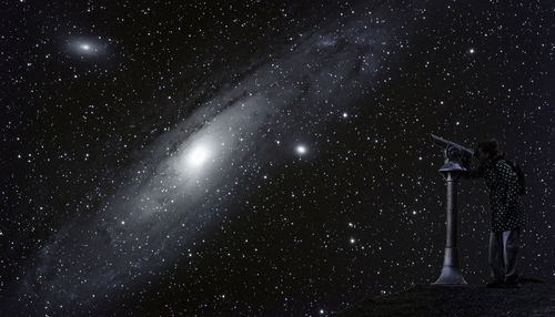 Woman looking through telescope while standing against sky at night