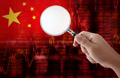 Digital composite image of cropped hand holding magnifying glass against chinese flag
