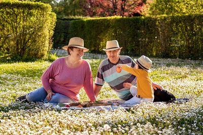 Happy senior family wearing straw summer hats having picnic with little grandson on blooming meadow