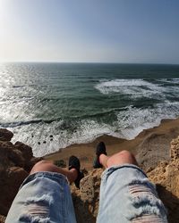 Low section of man sitting on rock by sea against sky