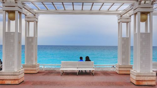 Rear view of people sitting on bench at pier while looking at horizon