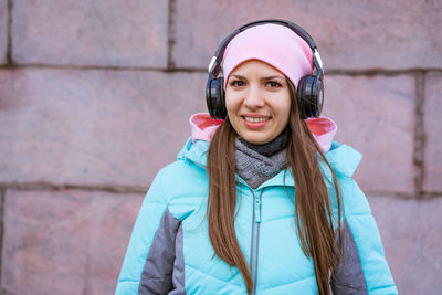 Happy young woman in warm jacket and pink hat listening to music on headphones