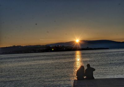 People sitting on shore against sky during sunset