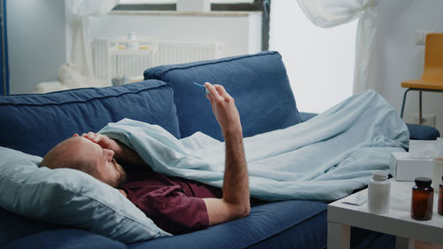 Sick man checking temperature while lying on sofa