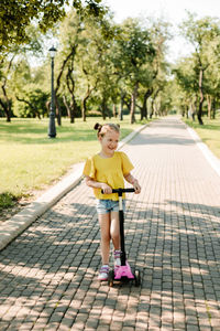 An active little girl rides a scooter on a path in an outdoor park on a summer day. 