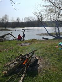 People relaxing on log by lake against sky